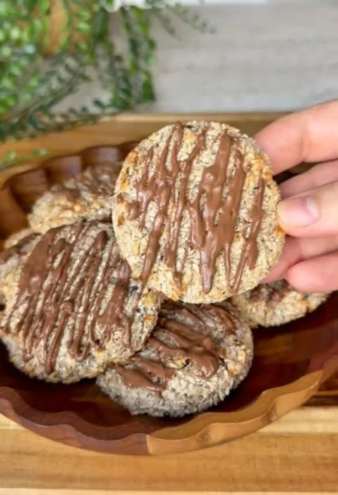 A hand holding a 2-ingredient banana coconut cookie drizzled with melted chocolate above a wooden bowl of more cookies