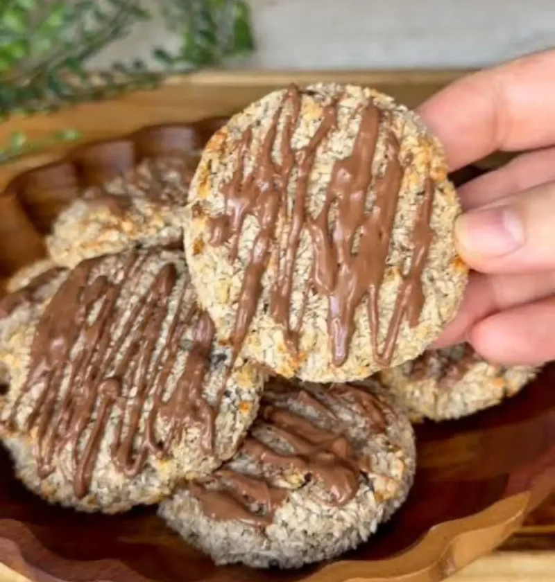 A hand holding a 2-ingredient banana coconut cookie drizzled with melted chocolate above a wooden bowl of more cookies