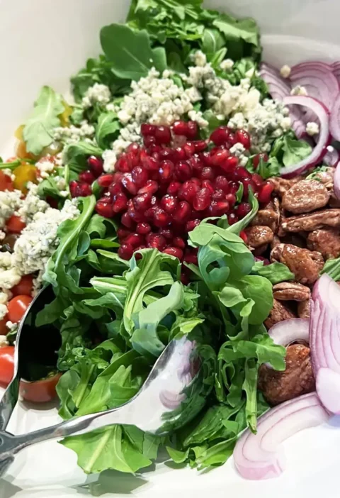 Arugula pomegranate salad in a white bowl with pecans, blue cheese, red onion and rainbow tomatoes