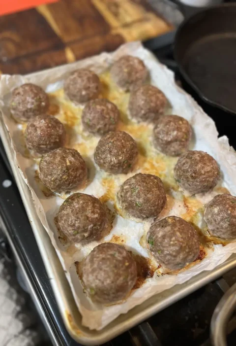 Baked Italian meatballs browned on a parchment-lined baking sheet next to a cast iron skillet