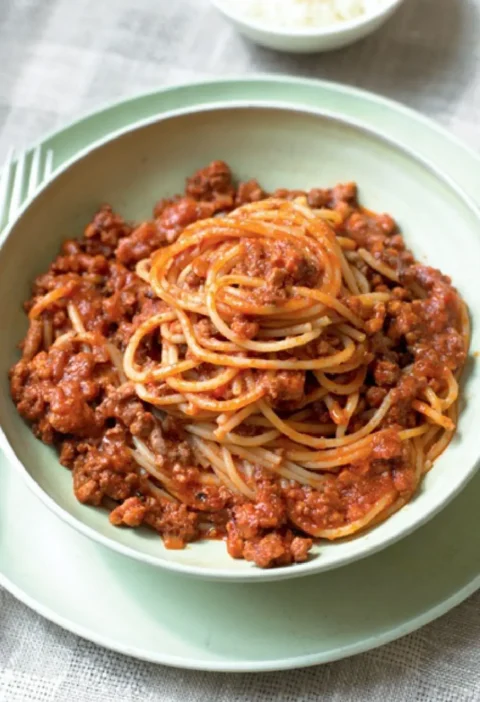 Bolognese sauce over spaghetti in a pale green bowl with a small bowl of grated cheese behind