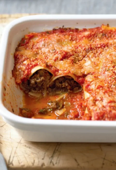 Baked cannelloni in a white dish with tomato sauce and Parmesan, one portion removed to show the beef filling