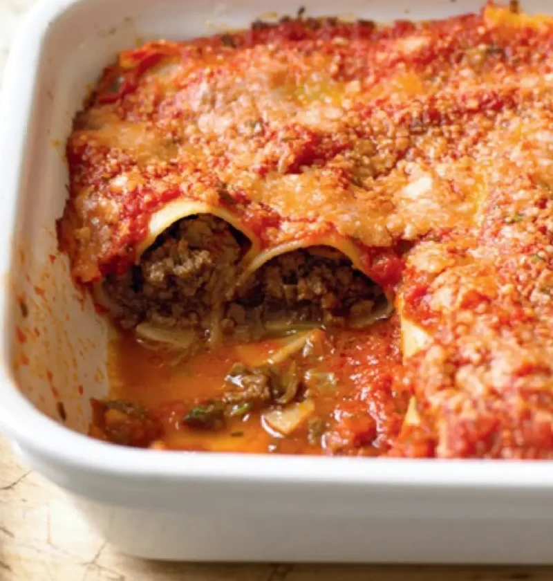 Baked cannelloni in a white dish with tomato sauce and Parmesan, one portion removed to show the beef filling
