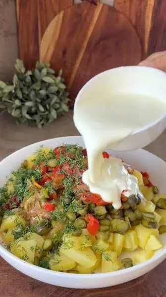 Yogurt dressing poured over diced potatoes, cornichons, roasted red pepper and dill in a white bowl