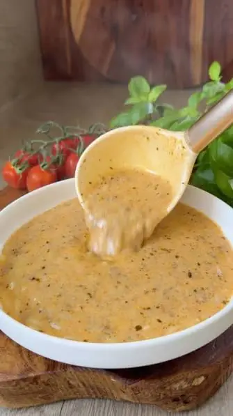 A ladle lifting creamy spinach soup from a white bowl with fresh basil and cherry tomatoes behind