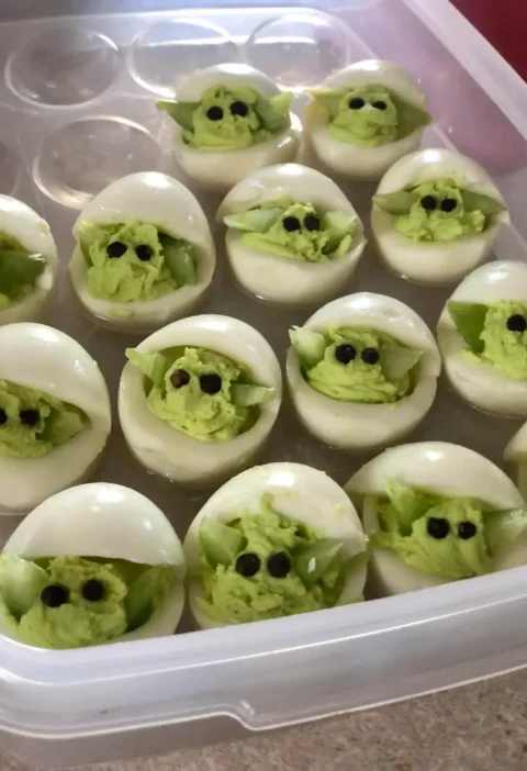 Deviled eggs Baby Yoda with green filling, peppercorn eyes and celery ears in a plastic egg carrier