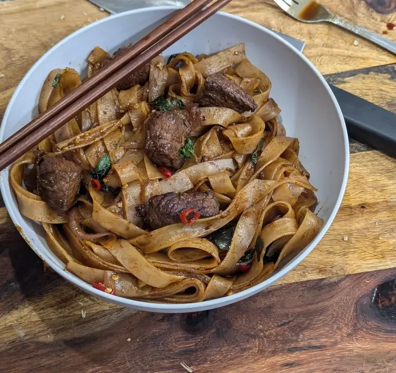 Bowl of Italian drunken noodles with seared beef, sliced red chilies and basil, with chopsticks resting on the rim