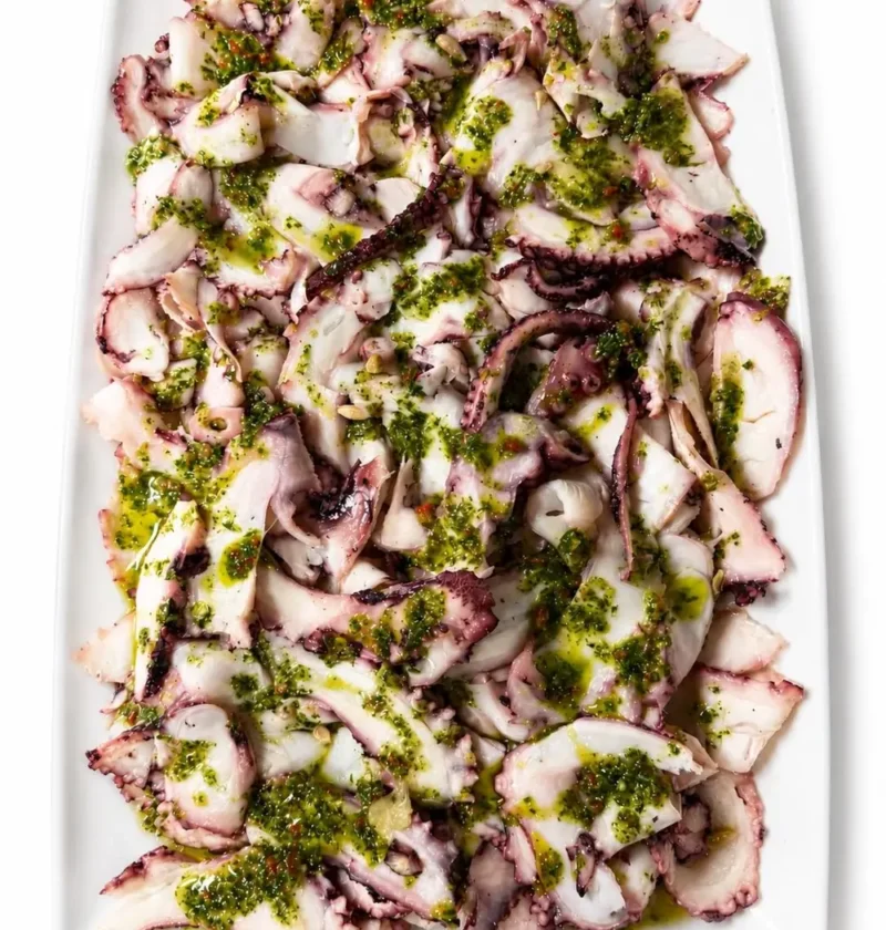 Octopus carpaccio thin slices on a white rectangular platter topped with green parsley and chili dressing