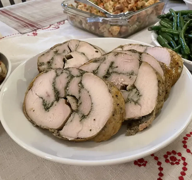 Sliced porchetta style turkey on a white plate showing herb pinwheels, with green beans and stuffing behind