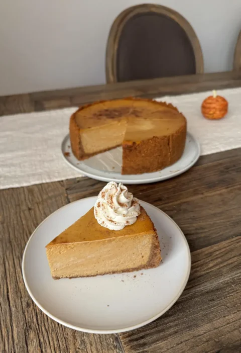 A slice of pumpkin cheesecake with whipped cream in front of the whole cheesecake on a wooden table