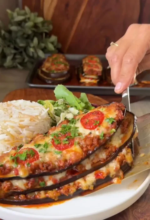 A spatula lifting an eggplant tower with beef, melted cheese and tomato slices from a plate with rice and salad