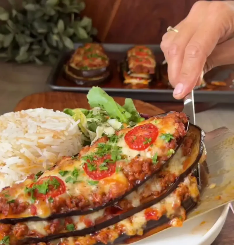 A spatula lifting an eggplant tower with beef, melted cheese and tomato slices from a plate with rice and salad