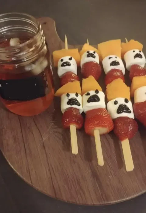 Halloween ghost fruit skewers with strawberries, marshmallow ghosts and cantaloupe on a wooden board next to a jar of punch