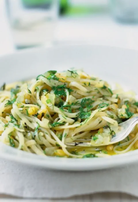 Herb sauce tossed with linguine in a white bowl, with chopped green herbs, onion and pine nuts