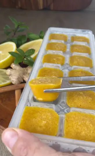 Ginger turmeric tea cubes in an ice cube tray, one cube lifted out with tongs, with lemon and ginger behind