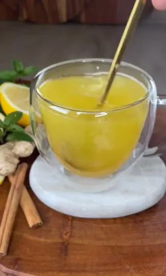 Immune-boosting ginger turmeric tea in a glass cup, stirred with a gold spoon, with lemon, ginger and cinnamon behind