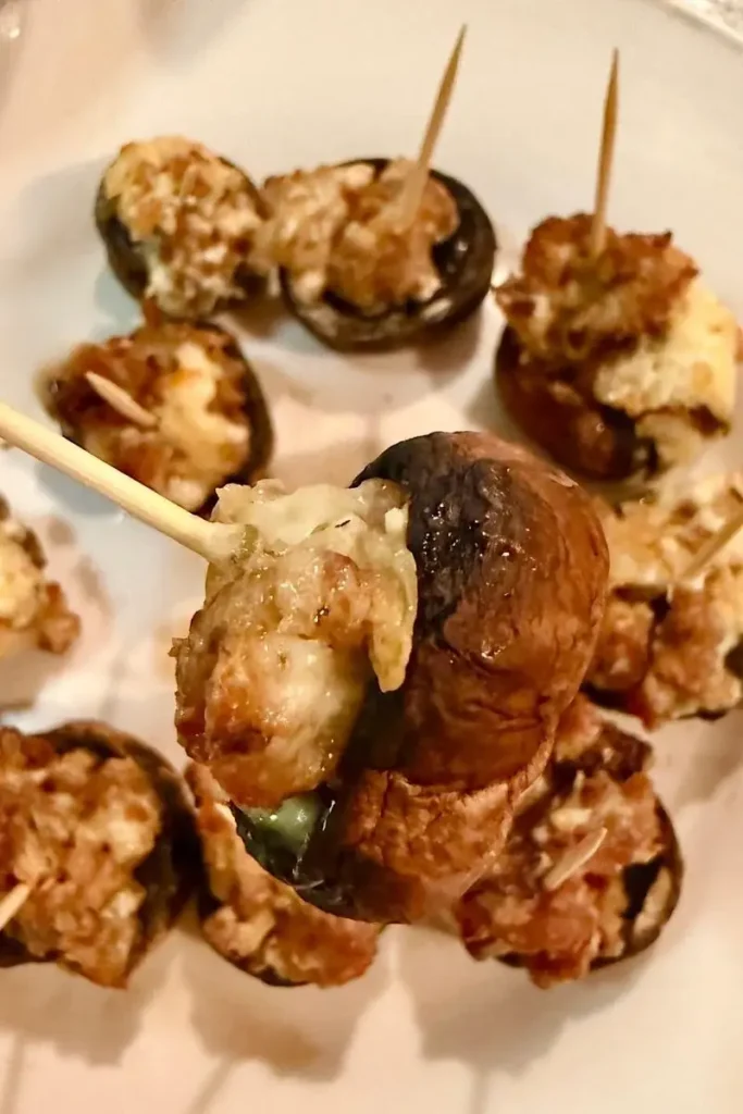 Italian stuffed mushrooms with sausage and cheese filling held up on a toothpick above a plate of more stuffed caps