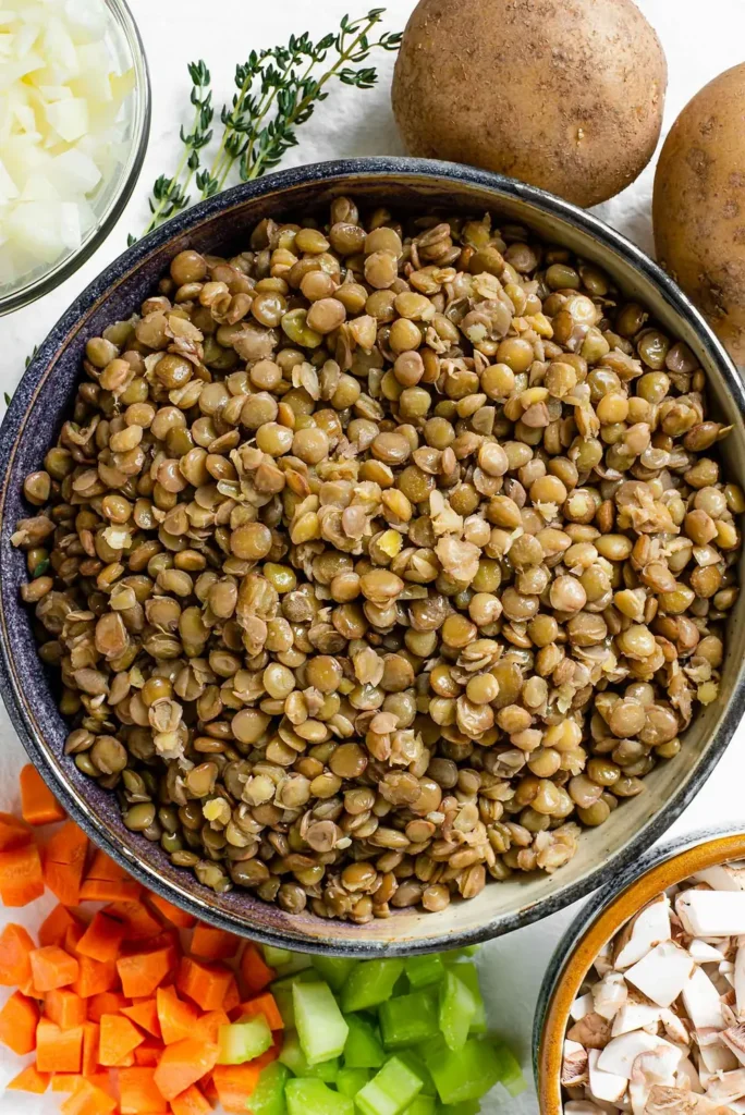 Cooked green lentils in a bowl with potatoes, diced carrots, celery, onion and mushrooms around it