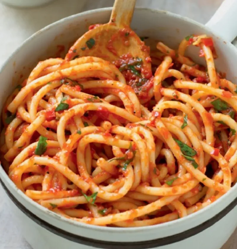 Neapolitan-style macaroni in a white pot with a wooden spoon, thick pasta strands coated in tomato sauce and parsley