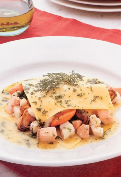 Open raviolo with fish: a pasta square over salmon, scallops and mussels in dill butter sauce on a white plate