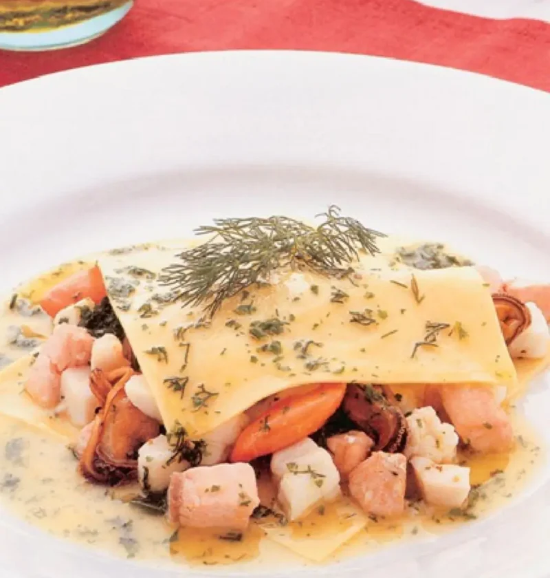 Open raviolo with fish: a pasta square over salmon, scallops and mussels in dill butter sauce on a white plate