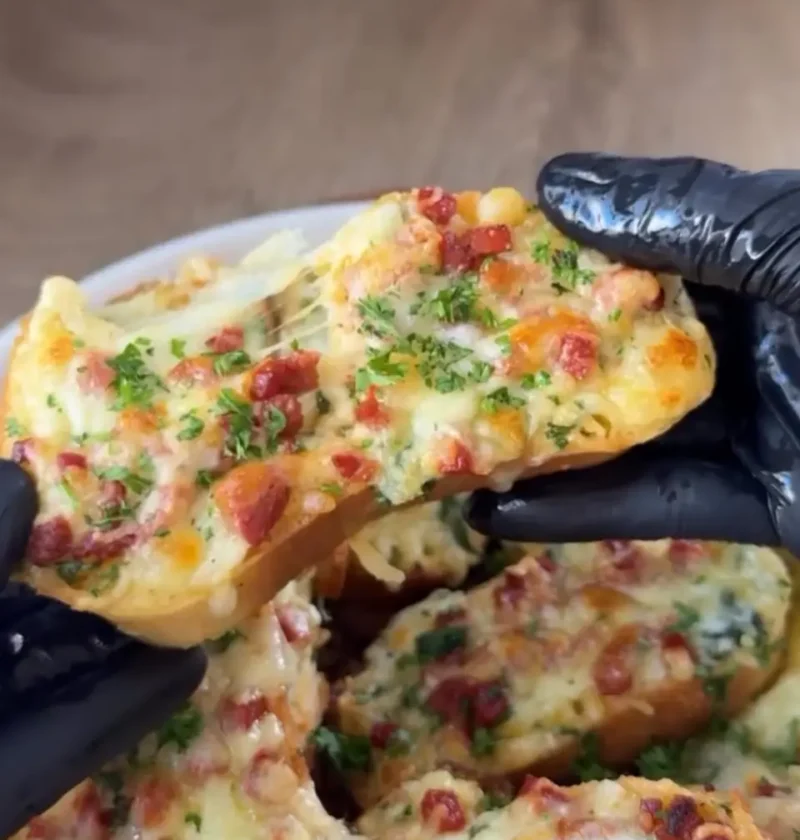 A hand in a black glove holding a cheesy egg toast slice topped with sucuk and parsley, with more slices behind it