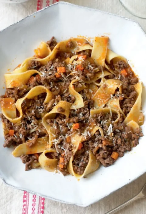 Pappardelle with meat sauce topped with grated Parmesan in a white octagonal bowl