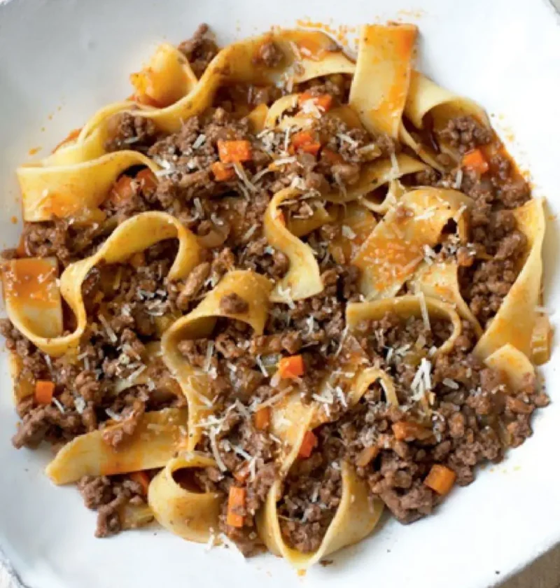 Pappardelle with meat sauce topped with grated Parmesan in a white octagonal bowl
