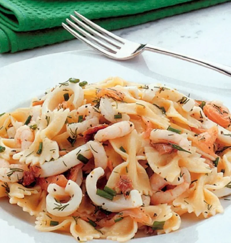 Pasta salad with fish on a white plate with farfalle, squid rings, shrimp, smoked salmon and fresh chives