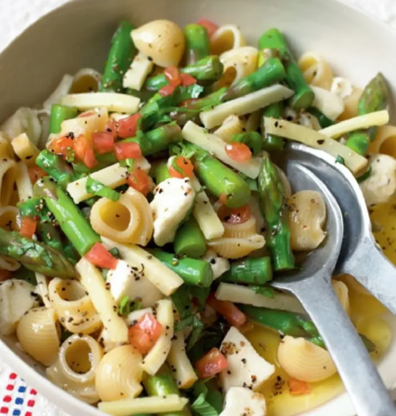 Pasta salad with vegetables in a white bowl with gomiti, asparagus, celeriac, tomato and mozzarella, and serving spoons