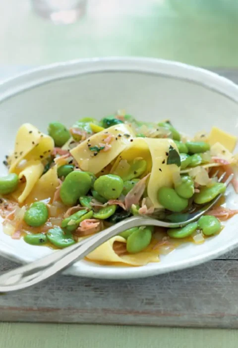 A white bowl of pappardelle with green beans, ham strips and basil, with a fork resting in it