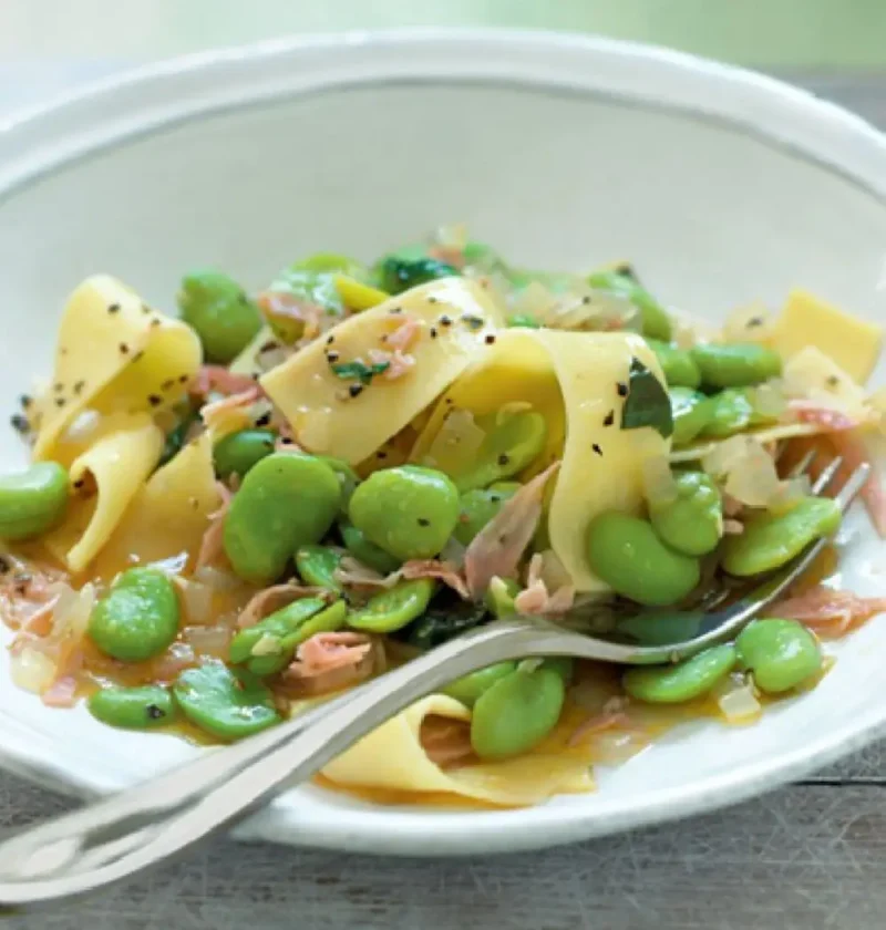 A white bowl of pappardelle with green beans, ham strips and basil, with a fork resting in it