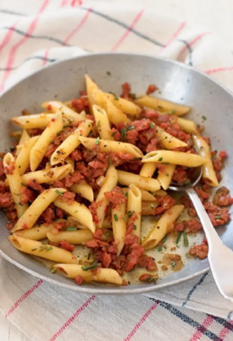 Penne rigate with sausage crumbles and chopped rosemary in a shallow metal pan with a spoon