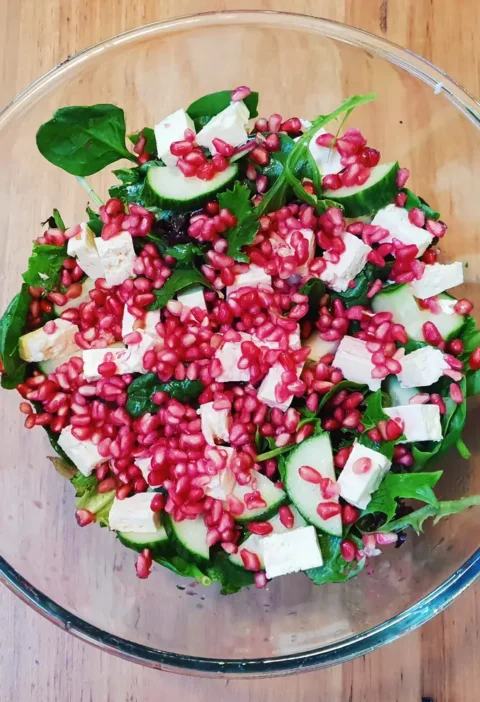 Pomegranate salad in a glass bowl with greens, cubed feta, cucumber slices and pomegranate seeds