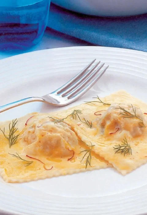 Two ravioloni with fish on a white plate with saffron threads, dill sprigs and a fork