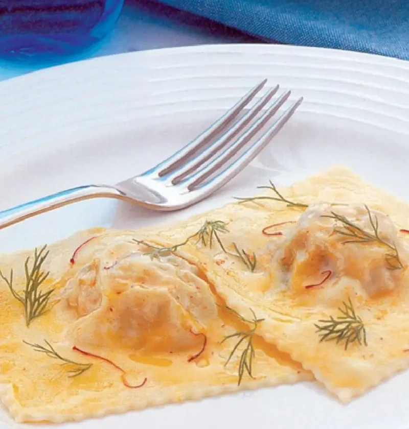 Two ravioloni with fish on a white plate with saffron threads, dill sprigs and a fork