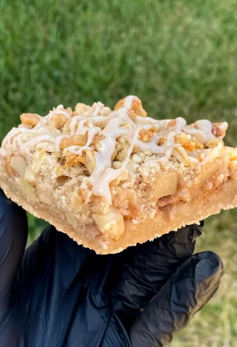 A ricotta apple bar with oat crumb topping and vanilla glaze held in a gloved hand