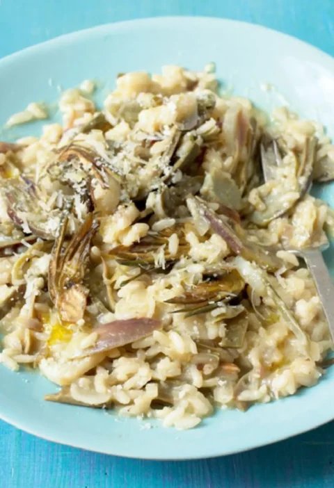 Risotto with two artichokes in a pale blue bowl, topped with slices of artichoke and grated Parmesan