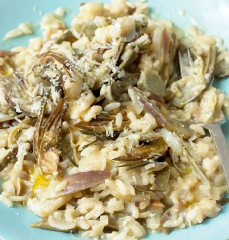 Risotto with two artichokes in a pale blue bowl, topped with slices of artichoke and grated Parmesan