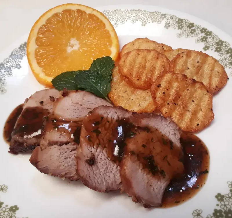 Sliced roasted pork with orange sauce on a plate with an orange half, mint and crinkle-cut potatoes