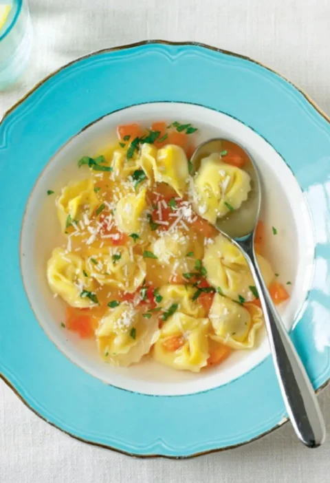 Soup with cappelletti in a blue bowl with diced tomato, parsley and grated Parmesan