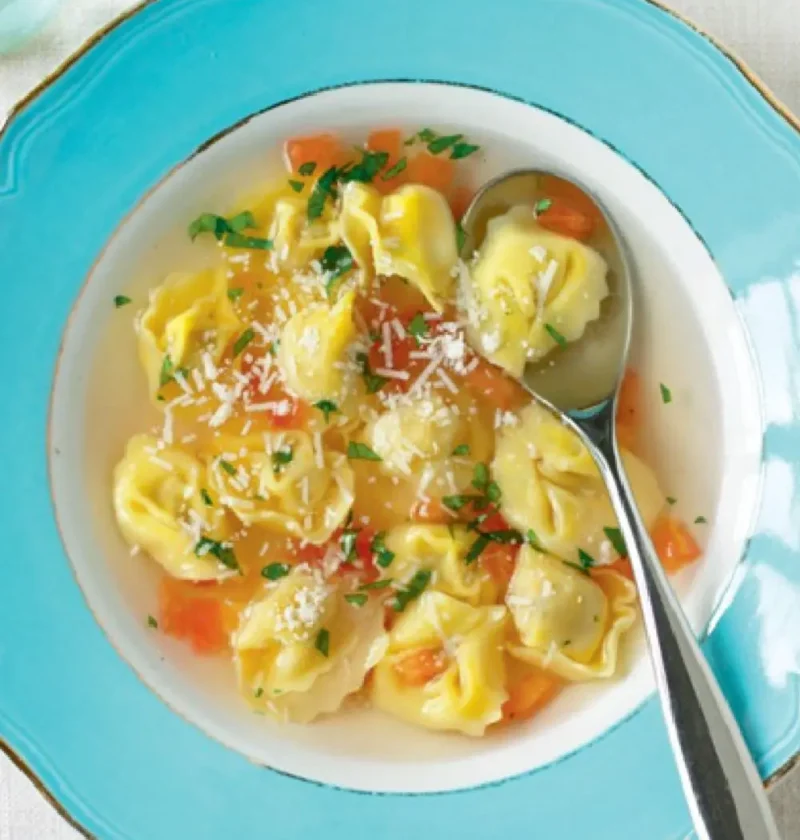 Soup with cappelletti in a blue bowl with diced tomato, parsley and grated Parmesan