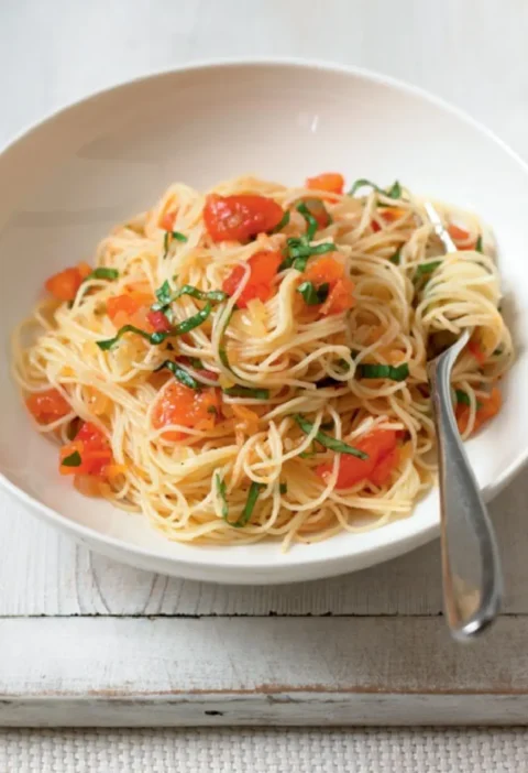 Spaghettini with tomato and basil in a white bowl, topped with chopped tomato and shredded basil, with a fork