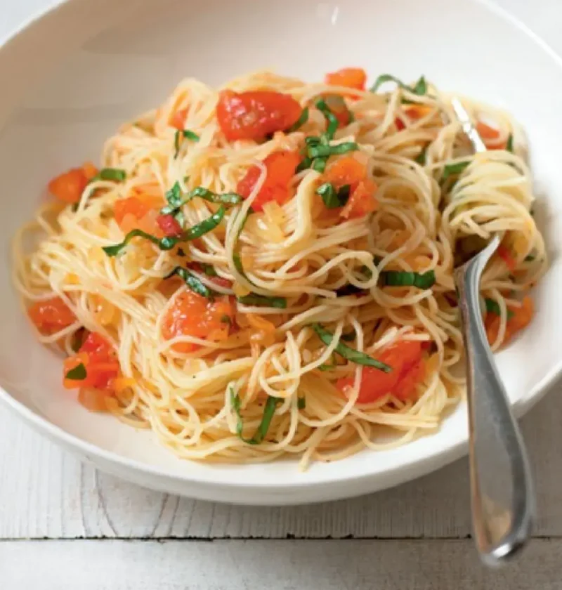 Spaghettini with tomato and basil in a white bowl, topped with chopped tomato and shredded basil, with a fork
