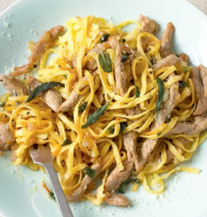 Tagliolini with veal strips and sage leaves on a pale plate with a fork and grated Parmesan nearby